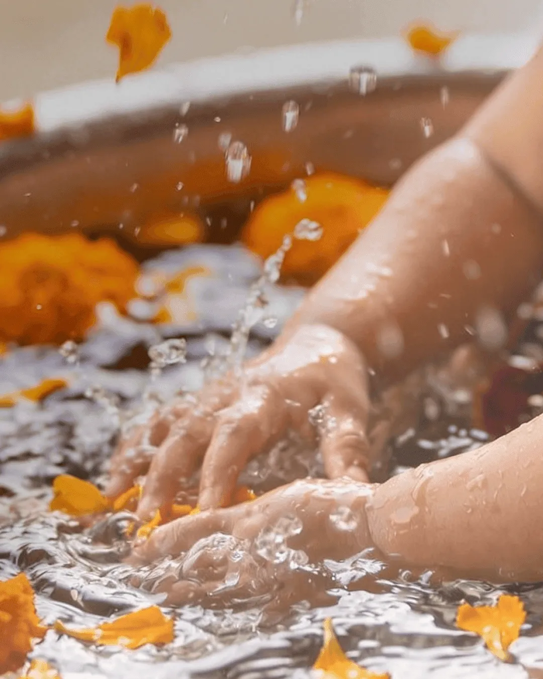 A child's hands playing in bath water strewn with marigold petals during an ubtan bath