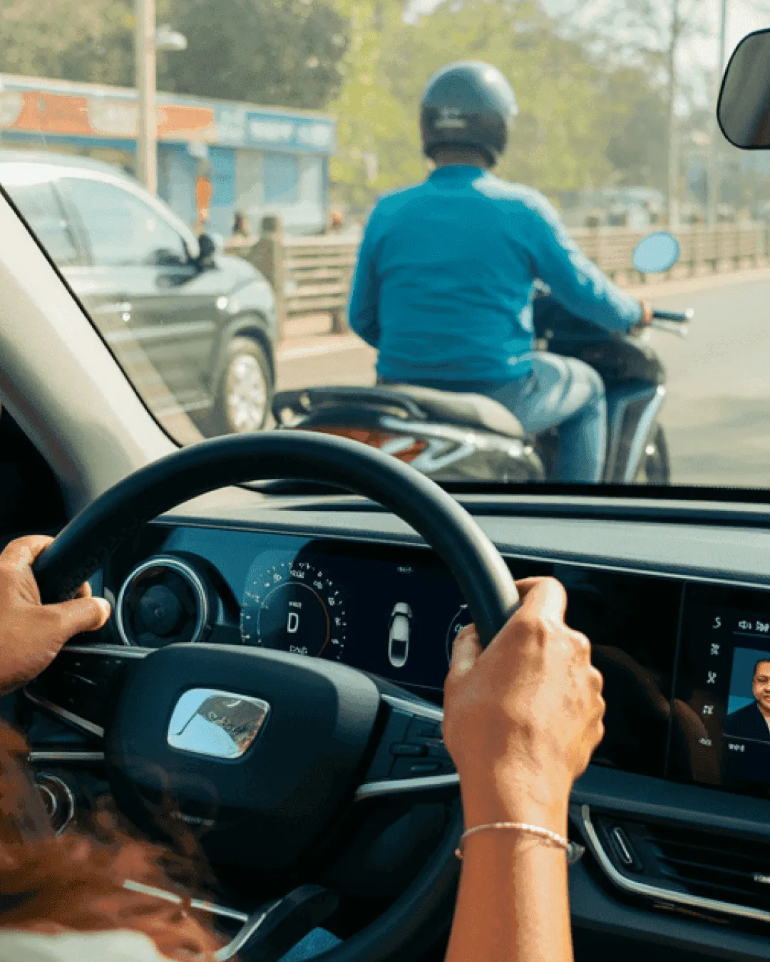 A woman's hands on the steering wheel of a car, driving through a Delhi street with a motorcyclist ahead