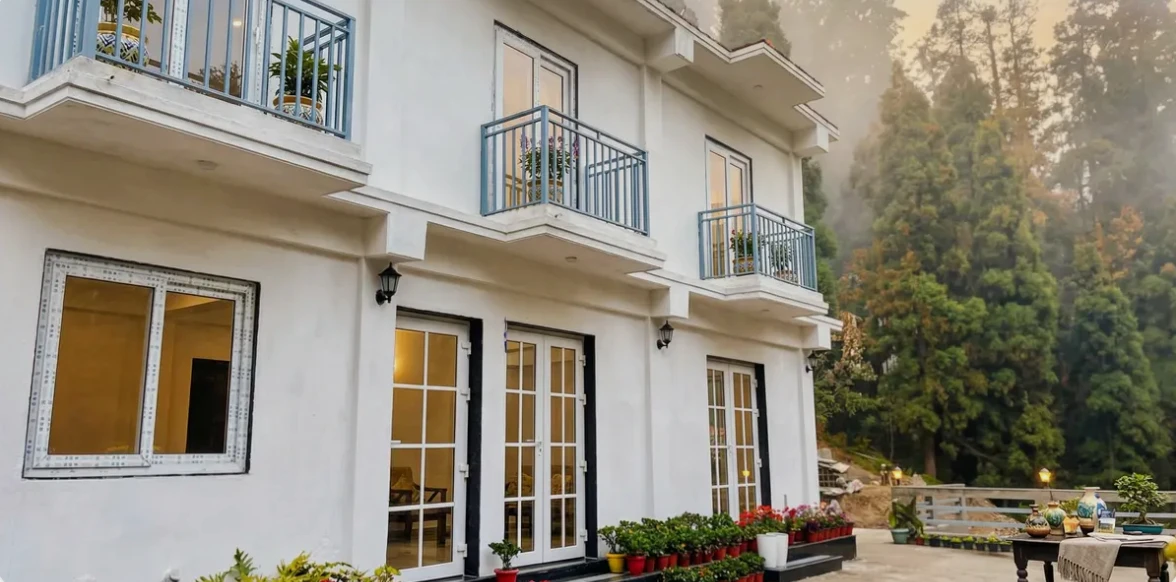 Exterior of Kulam Homestay in Mirik, a white hillside house with blue balcony railings, garden plants and forest behind